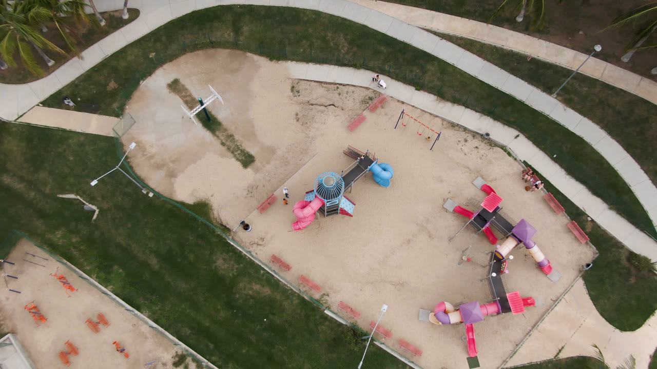 Aerial frame from San Jose del Cabo with the playground shifted slightly in view, showing the blue slide tower, pink slide, curved paths, benches, and open sand.