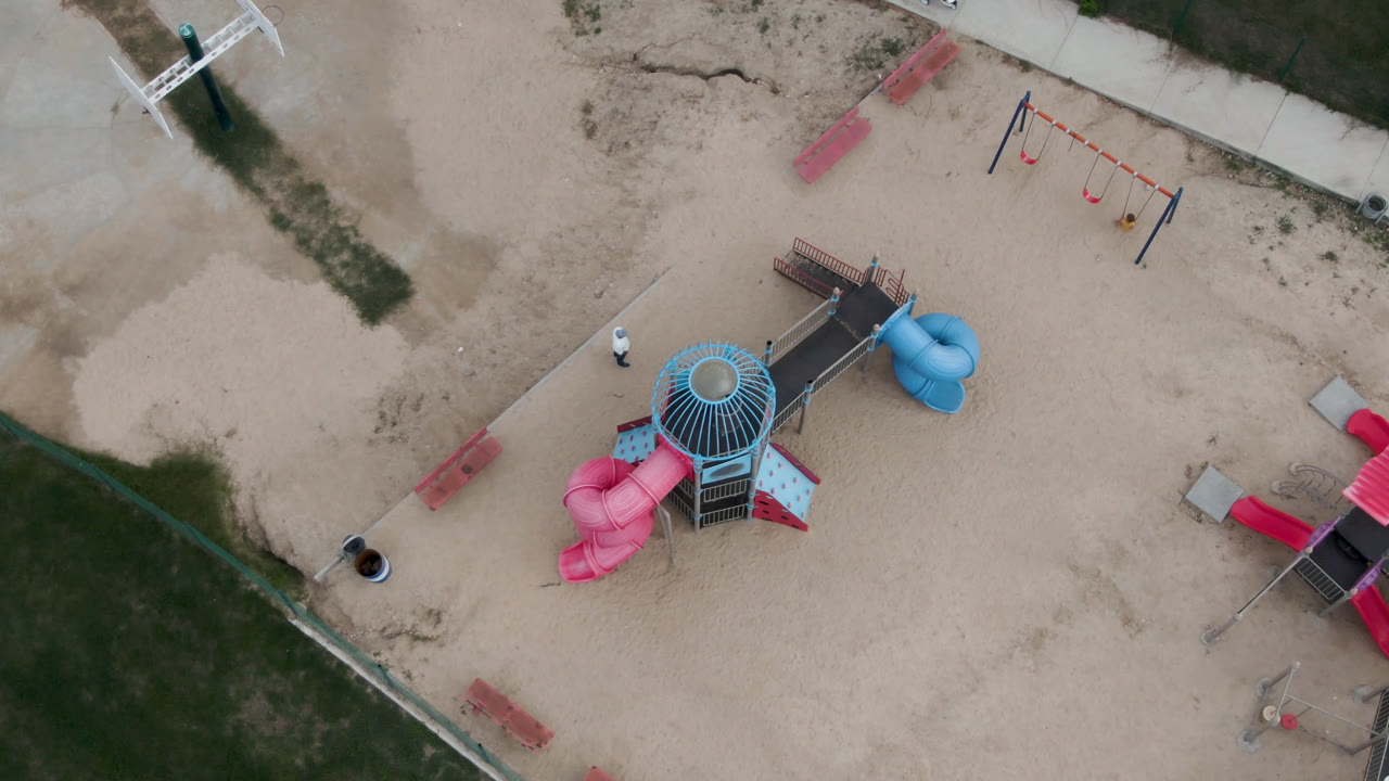 Top-down view of a sandy playground with pink and blue slides and nearby paths.
