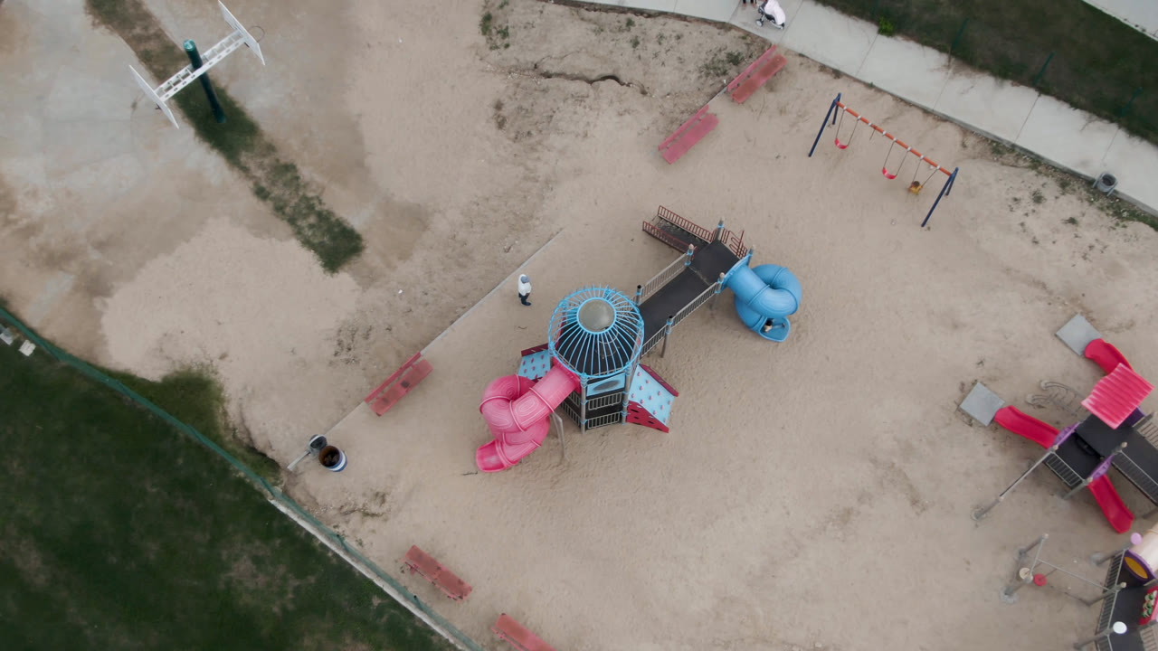 High aerial frame from San Jose Del Cabo with the central playground tower, pink and blue slides, a swing set, benches, and curved walkways visible around the sand.