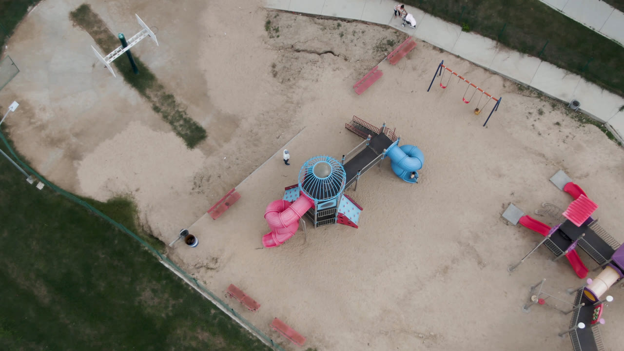 Overhead San Jose Del Cabo frame showing the playground split between a central slide tower and a red-roofed play structure, with swings, benches, sand, paths, and grass in view.