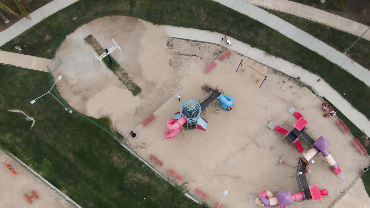 Overhead frame of a sand playground in San Jose del Cabo with pink slides, a blue play piece, grass borders, curved paths, and no visible people.