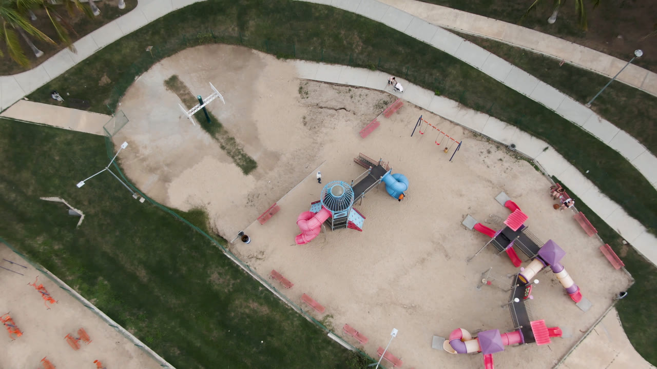 Top-down view in San Jose del Cabo showing the sand play area, pink slide cluster, blue equipment, purple tunnel section, surrounding lawn, and no visible people.