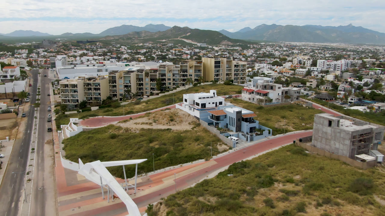 Aerial frame over San Jose del Cabo with the road running vertically near the left side, beige apartment buildings near center, white homes and scrubland below, distant mountains, and no visible people.