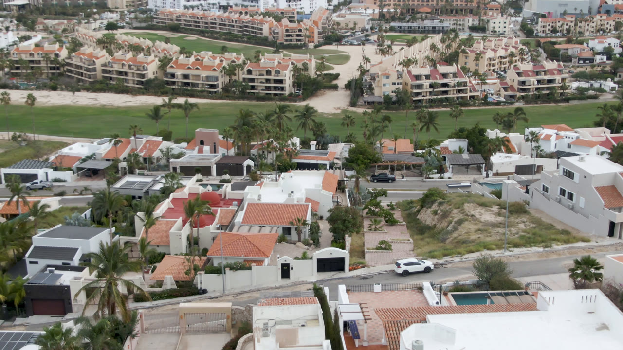 Aerial frame over San Jose del Cabo showing white residential buildings and terracotta roofs in front of a green golf course, resort-style buildings, and the ocean horizon, with no visible people.