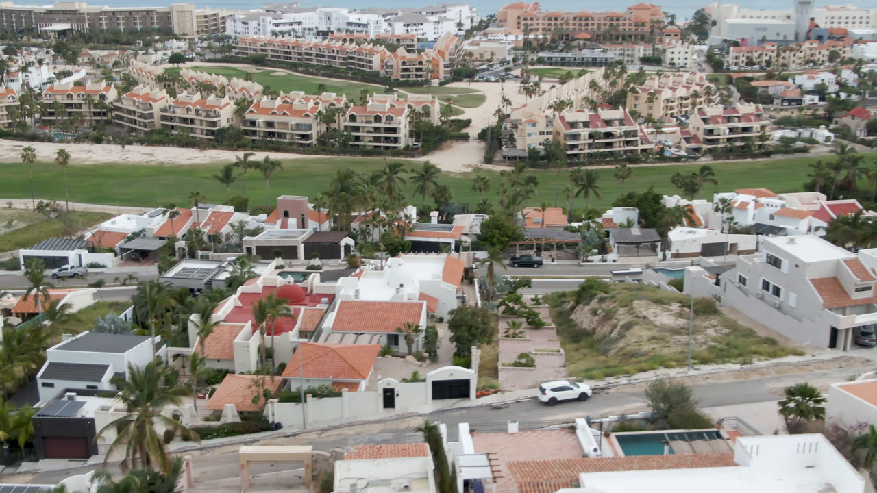 Drone-height view in San Jose del Cabo with white villas and a road across the lower frame, palm trees and golf-course grass in the center, and coastal buildings near the ocean, with no visible people.