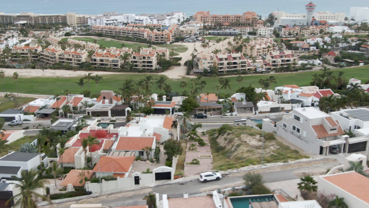 Wide aerial of residential rooftops, sandy lots, road traffic, golf course, and ocean.