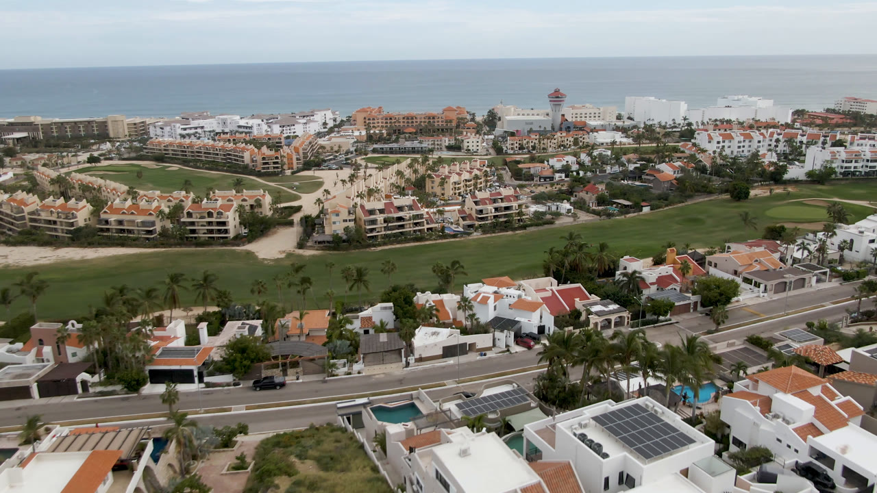 Aerial view in San Jose del Cabo with palm-lined residential streets at the bottom, a broad golf course through the center, large coastal hotels in back, blue ocean, and no visible people.
