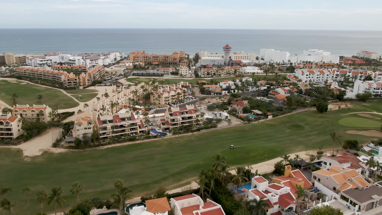 Second frame of the San Jose del Cabo aerial showing a wider stretch of fairway in the foreground, clustered resort buildings in the midground, ocean beyond, and no visible people.
