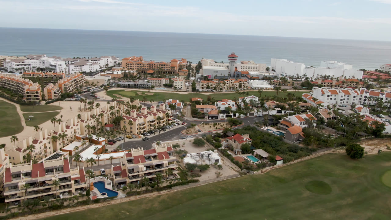 Second aerial frame in San Jose del Cabo showing more of the golf course edge, clustered white and terracotta buildings, beach strip, ocean, and no visible people.