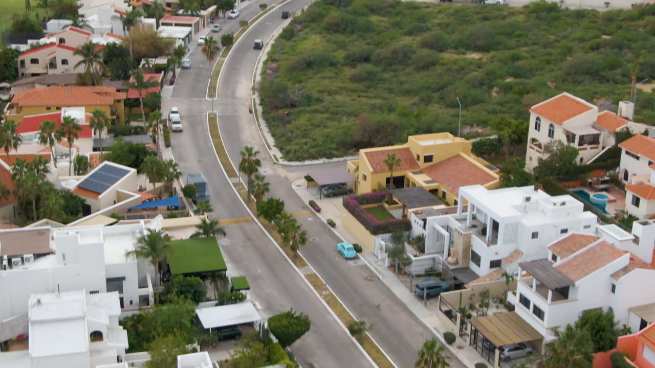 Aerial frame over San Jose del Cabo showing a curved residential road beside white and tan houses, parked cars, palm trees, and green open lots, with no visible people.