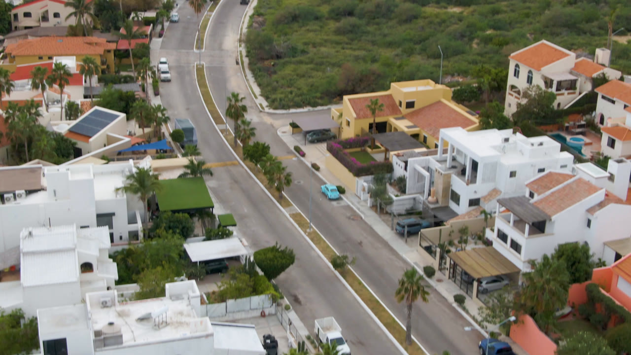 Elevated view in San Jose del Cabo with white stucco homes, red roof sections, a diagonal paved street, parked cars, palms, and undeveloped green land, with no visible people.