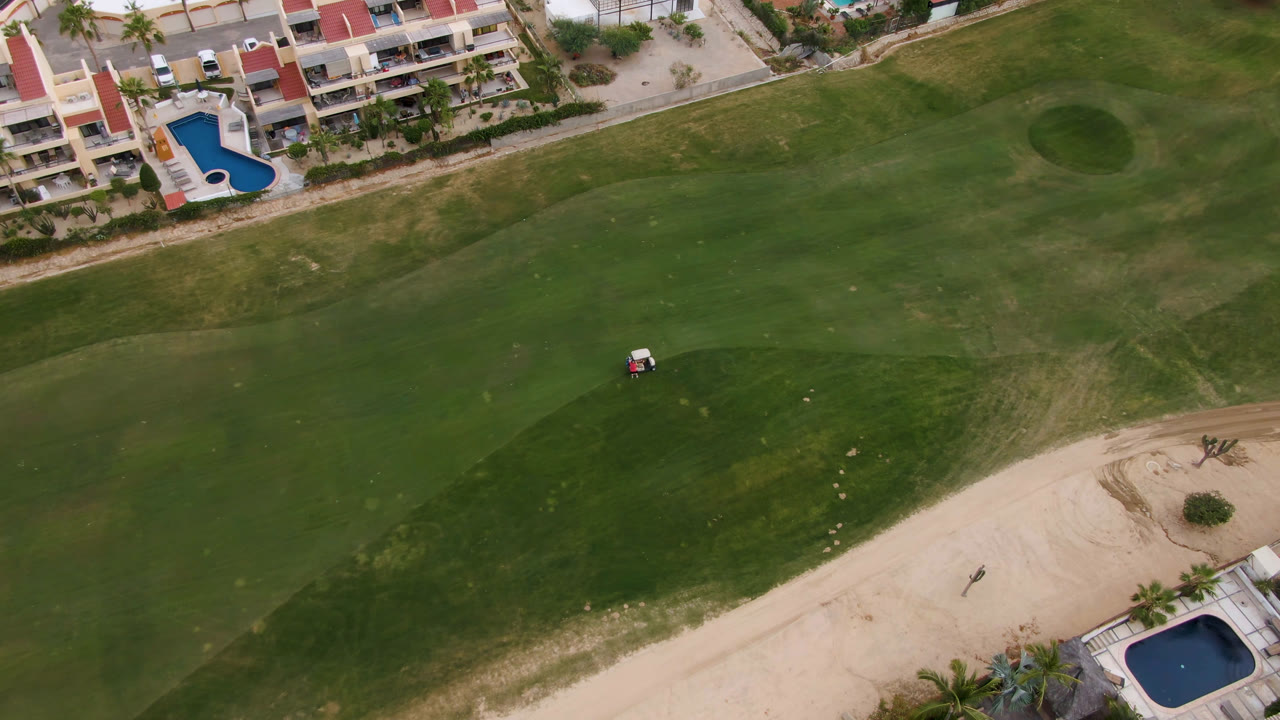 Overhead golf course with fairway, sand area, resort buildings, pools, and a small golf cart.