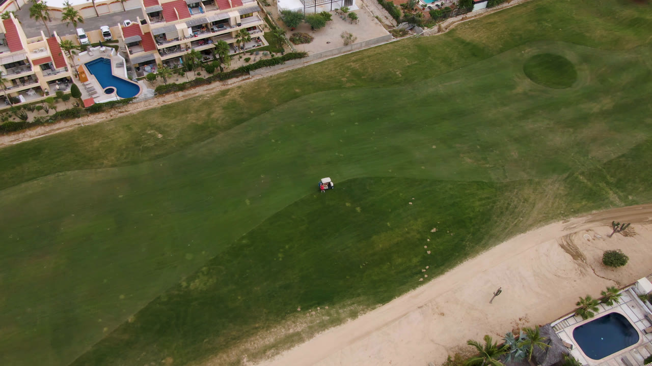 A second overhead frame from San Jose del Cabo shows the fairway stretching below resort buildings, with blue pools at the upper edge, a sand area at lower right, a small golf cart, and a distant figure.