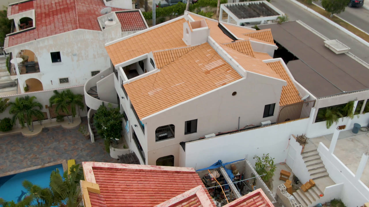 Elevated opening frame in San Jose del Cabo showing white stucco houses, red and orange tile roofs, small pools, palms, parked cars, and no visible people.