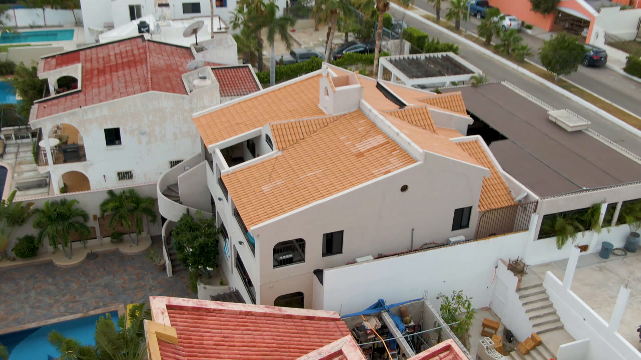Second frame in San Jose del Cabo with a large orange-tiled white house in the center, pools at the edges, palm landscaping, a paved road, and no visible people.