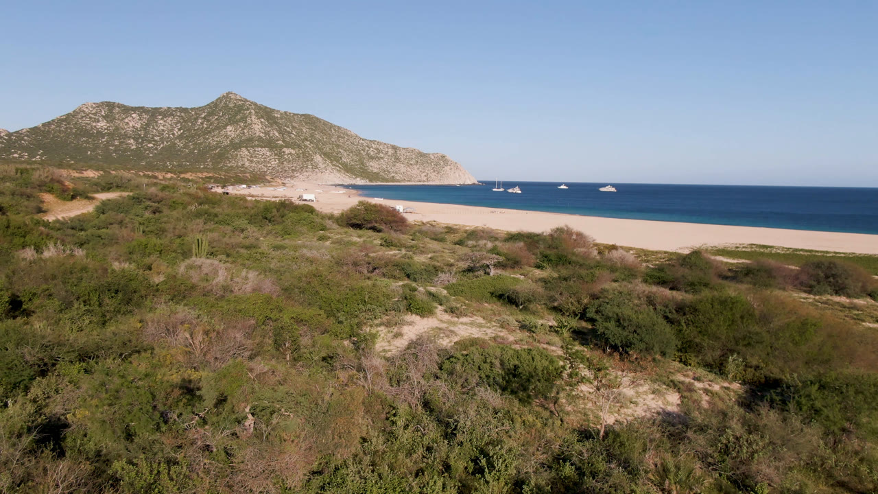 Wide beach, scrubland, blue ocean, boats, and rocky hills