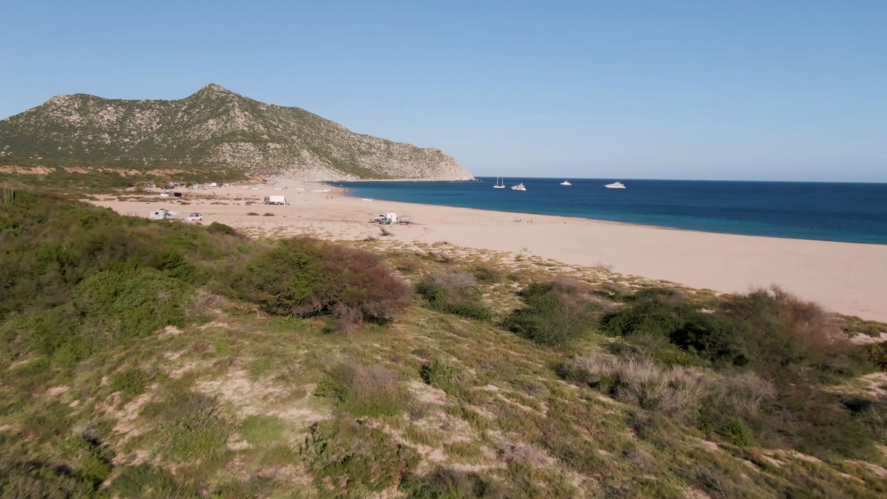 Wide view of Los Frailes, Baja California Sur, with scrubby dunes in front, a sandy beach, boats on blue water, rocky hills in back, and no visible people.