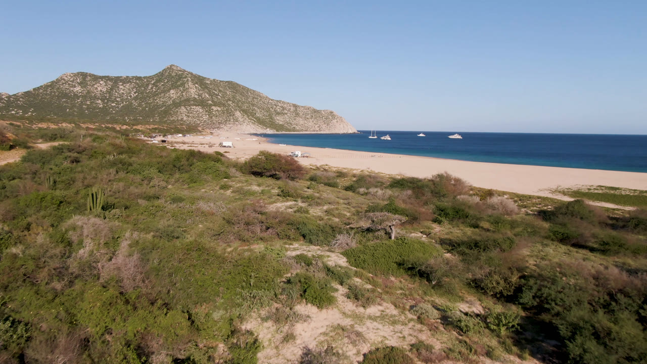 Wide aerial view at Los Frailes, Baja California Sur, Mexico, with scrubby foreground terrain, pale beach, scattered boats offshore, mountain ridges behind the bay, and no visible people.