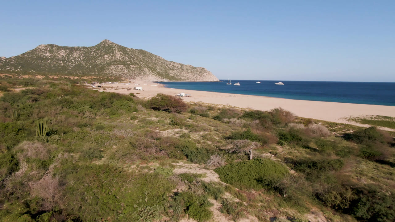 Aerial frame over Los Frailes with low green and brown scrub in front, a broad sandy beach, several boats on calm blue water, rocky hills in the distance, and no visible people.