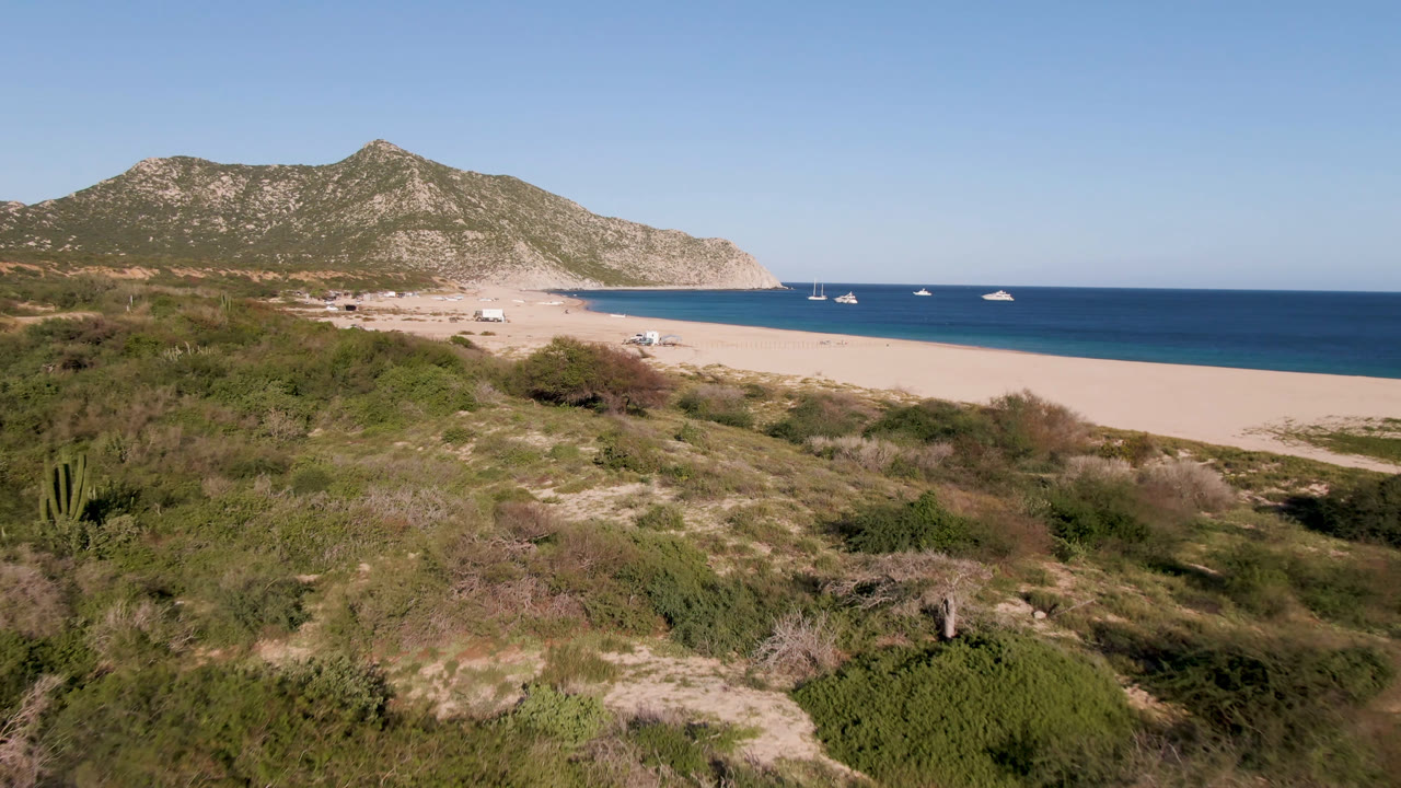Beach and bay below mountains with scattered boats and coastal brush in the foreground.