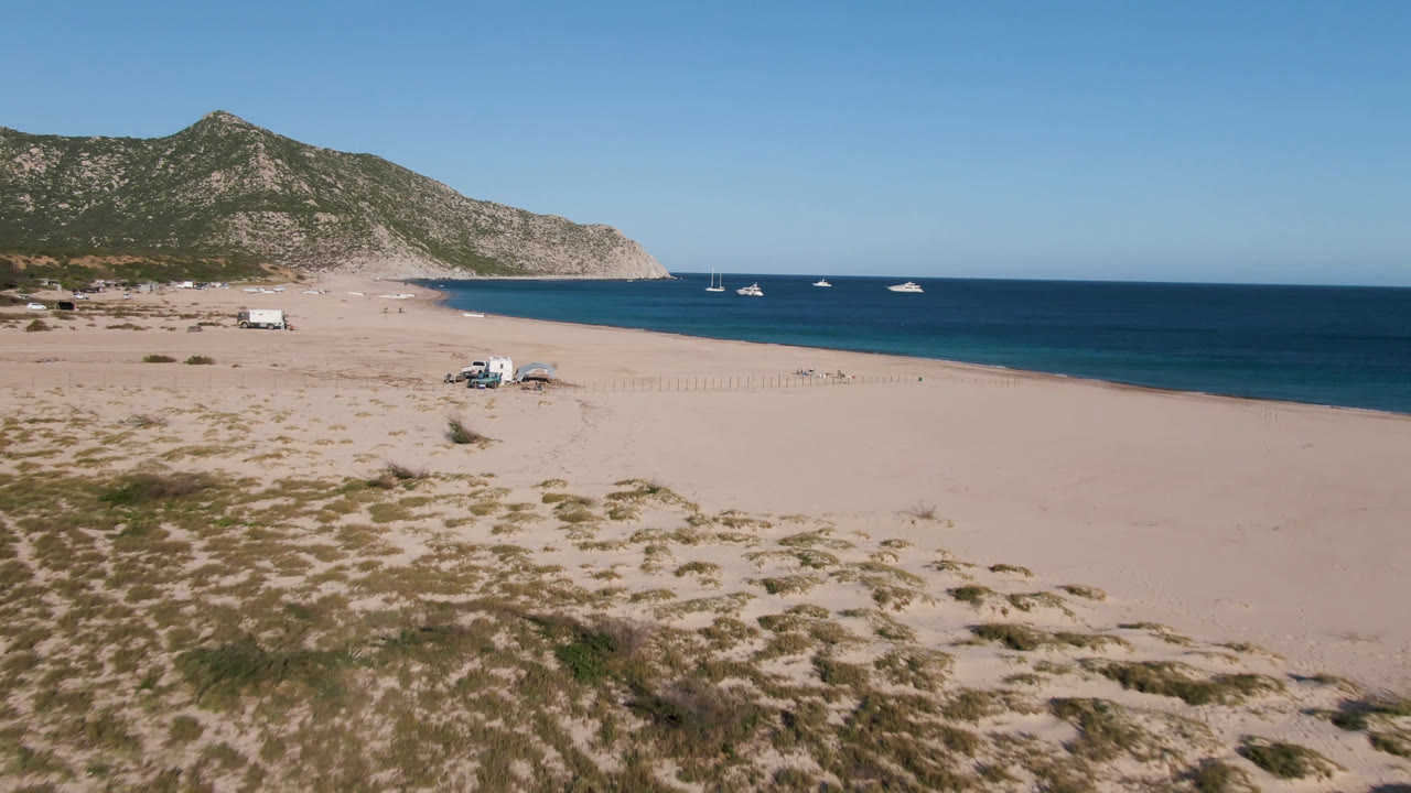 Opening aerial frame over Los Frailes, Baja California Sur, with scrubby sand in the foreground, a broad empty beach, anchored boats offshore, a rocky headland, and no visible people.