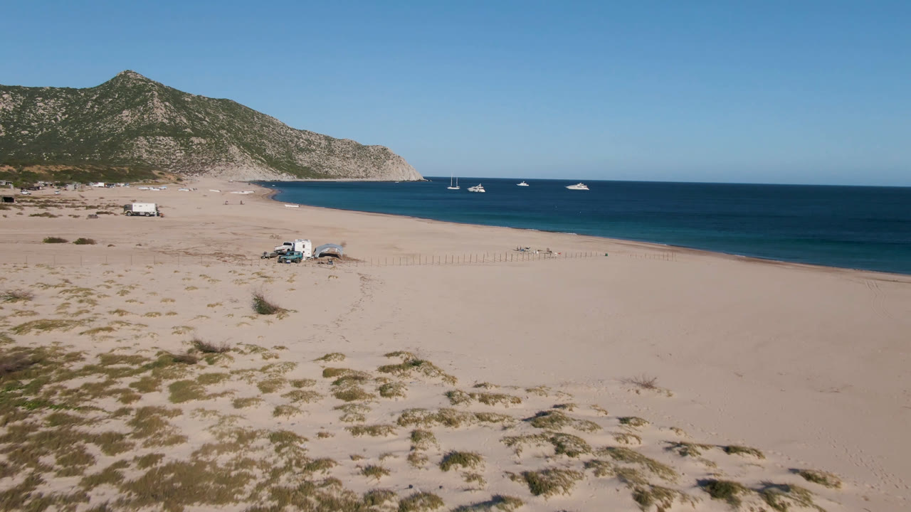 Los Frailes aerial view with a small camp setup on the sand below, anchored boats spread across the blue water, a long shoreline, rocky hills, and no visible people.