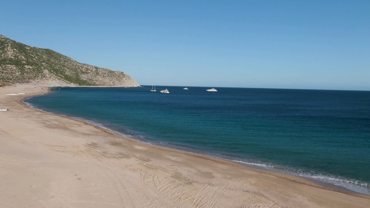Aerial view of Los Frailes with a wide sandy beach curving toward turquoise water, a rocky hillside at left, small boats offshore, and no visible people.