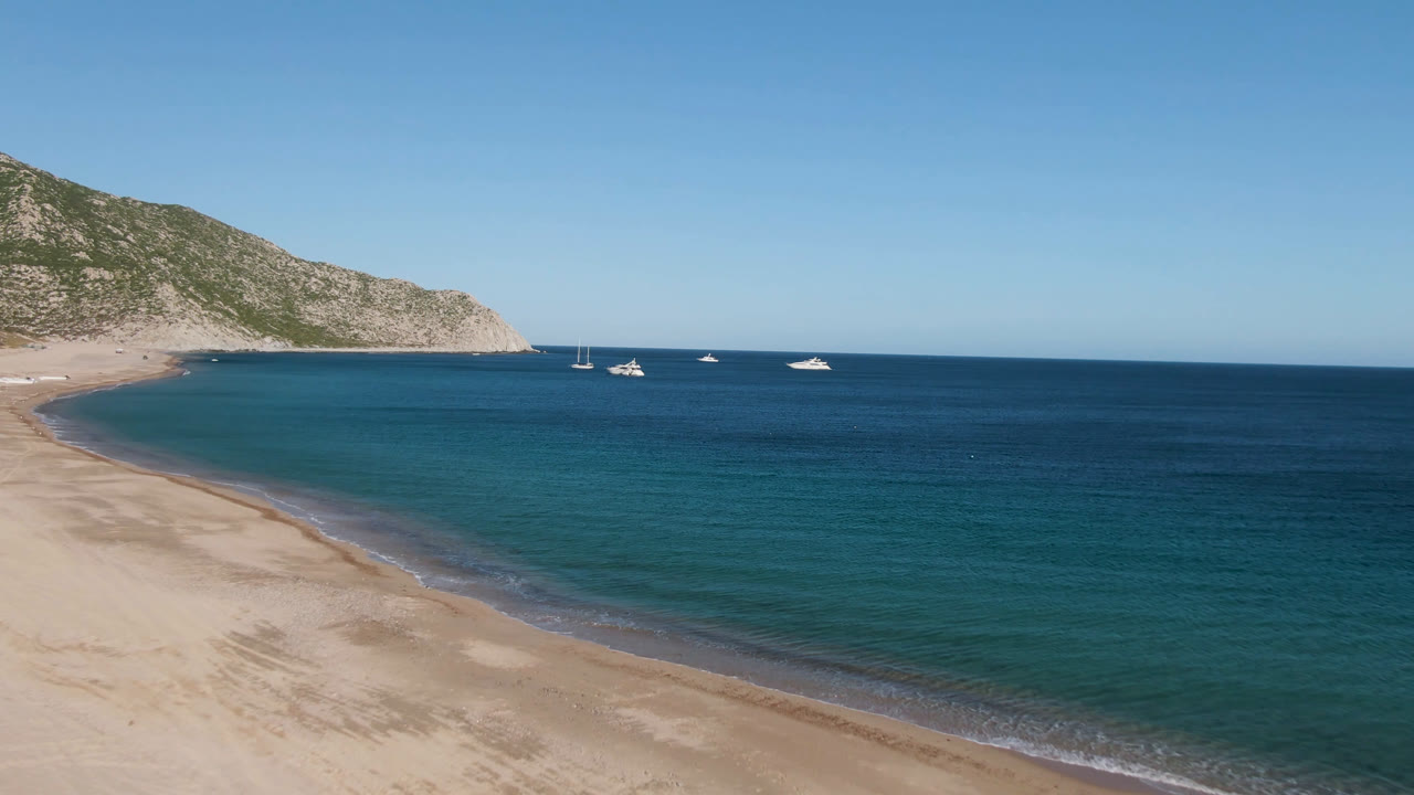 Aerial frame from Los Frailes with the beach lower left, calm blue-green shallows, distant white boats, a hillside headland, and no visible people.