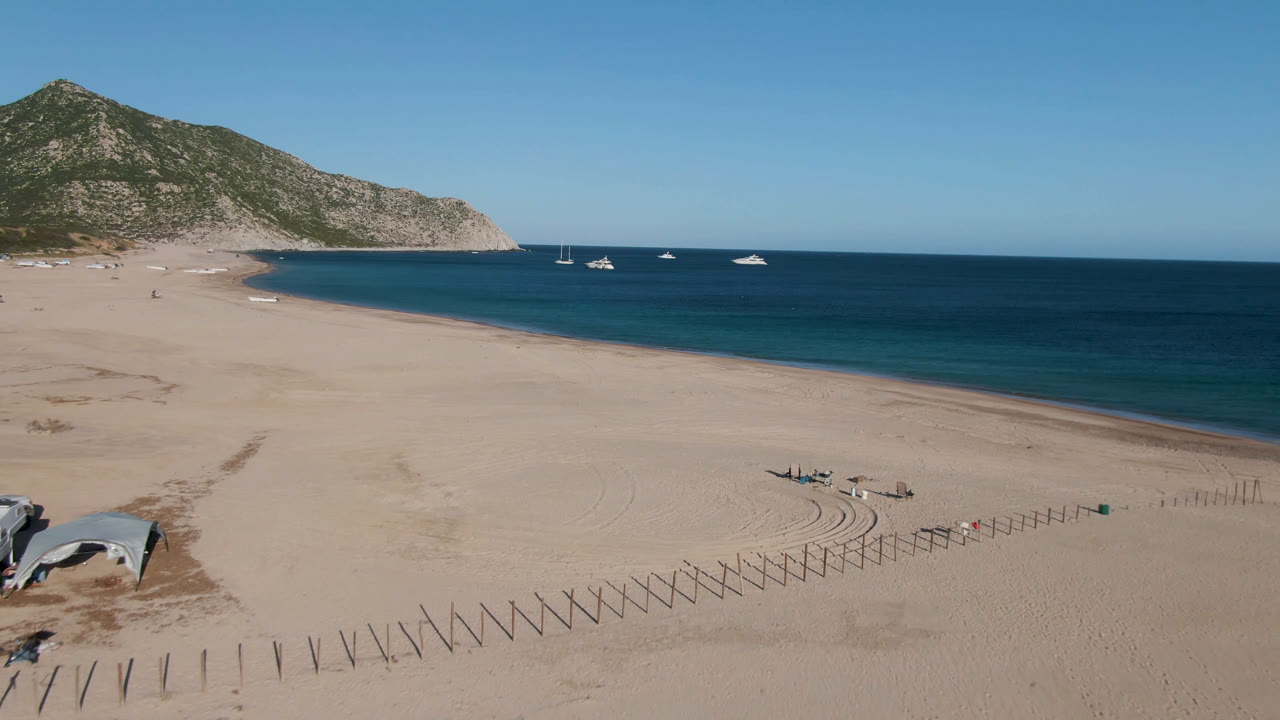 Aerial frame of Los Frailes, Baja California Sur with broad pale sand in front, several boats offshore, a rocky headland at left, and no visible people.