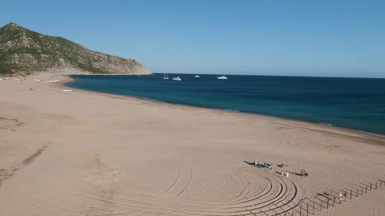 Drone frame over Los Frailes beach with sand tracks and low fencing in the lower frame, anchored boats across calm blue water, and no visible people.