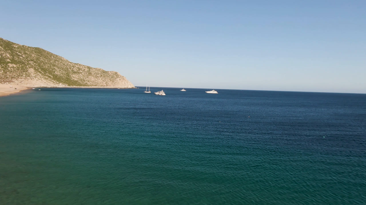 Aerial frame over Los Frailes, Baja California Sur, with blue-green water in front, small boats near the horizon, a rocky headland at left, and no visible people.