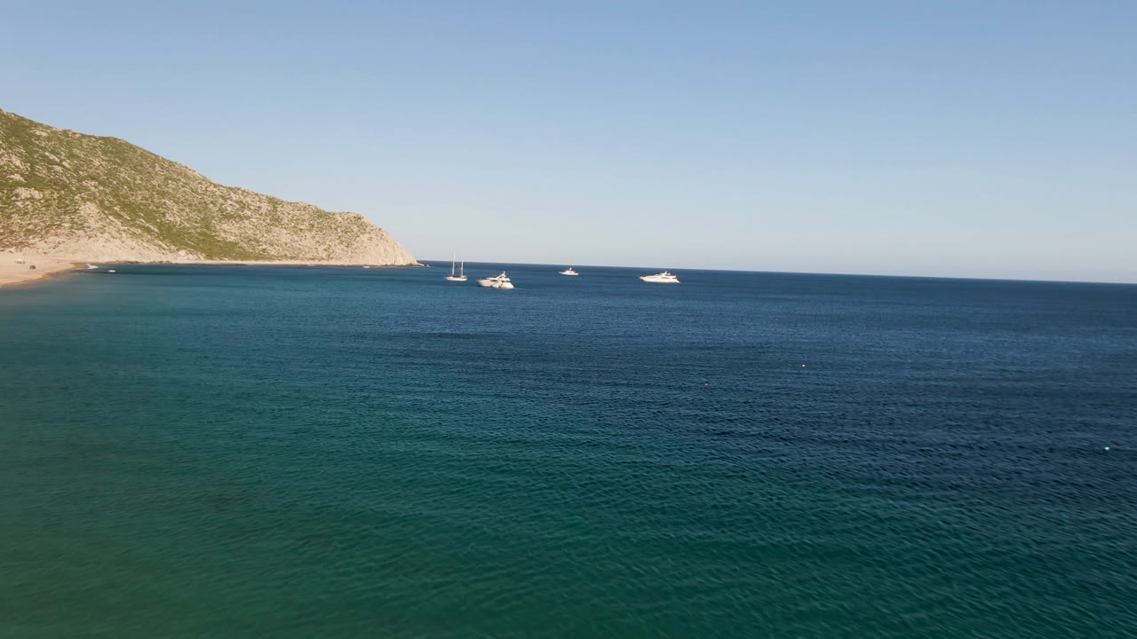 Second aerial frame at Los Frailes showing textured ocean surface, a line of small boats offshore, the rocky mountain on the left, and no visible people.