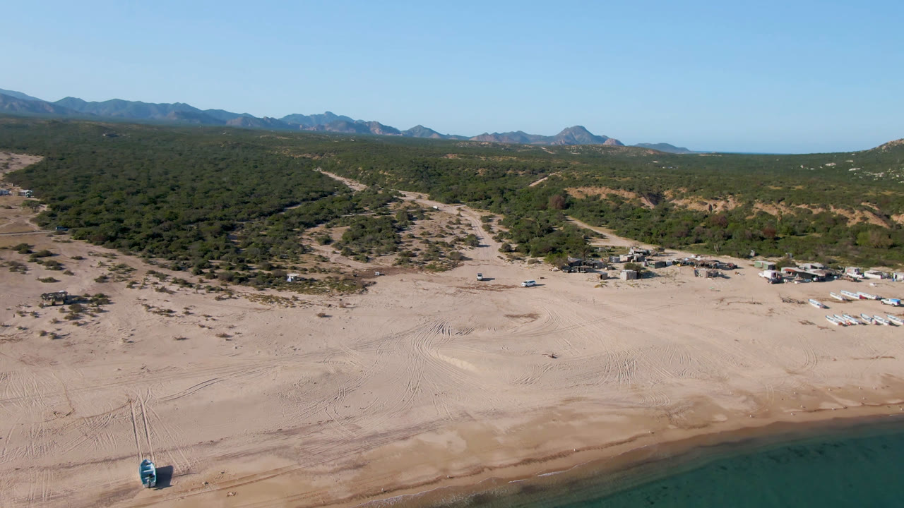 Wide aerial view over Los Frailes beach in Baja California Sur, with sandy foreground, blue-green water at the lower edge, tents near trees, distant mountains, and no visible people.