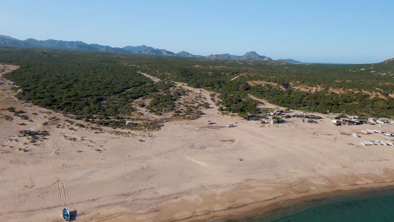 Los Frailes aerial frame showing the beach camp slightly farther along the shore, with pale sand, small tents or canopies, green scrubland, mountains on the horizon, and no visible people.