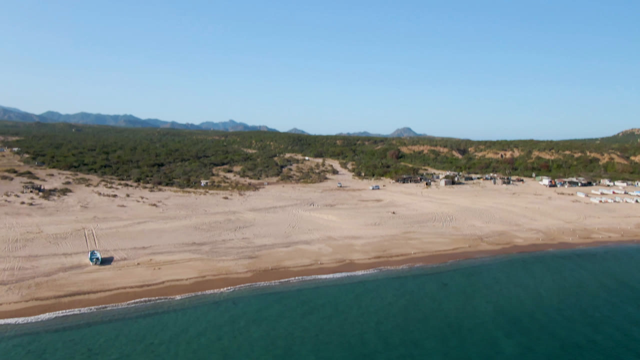 Opening aerial frame over Los Frailes, Baja California Sur, with blue water below a broad sand beach, green scrubland, distant mountains, and no visible people.