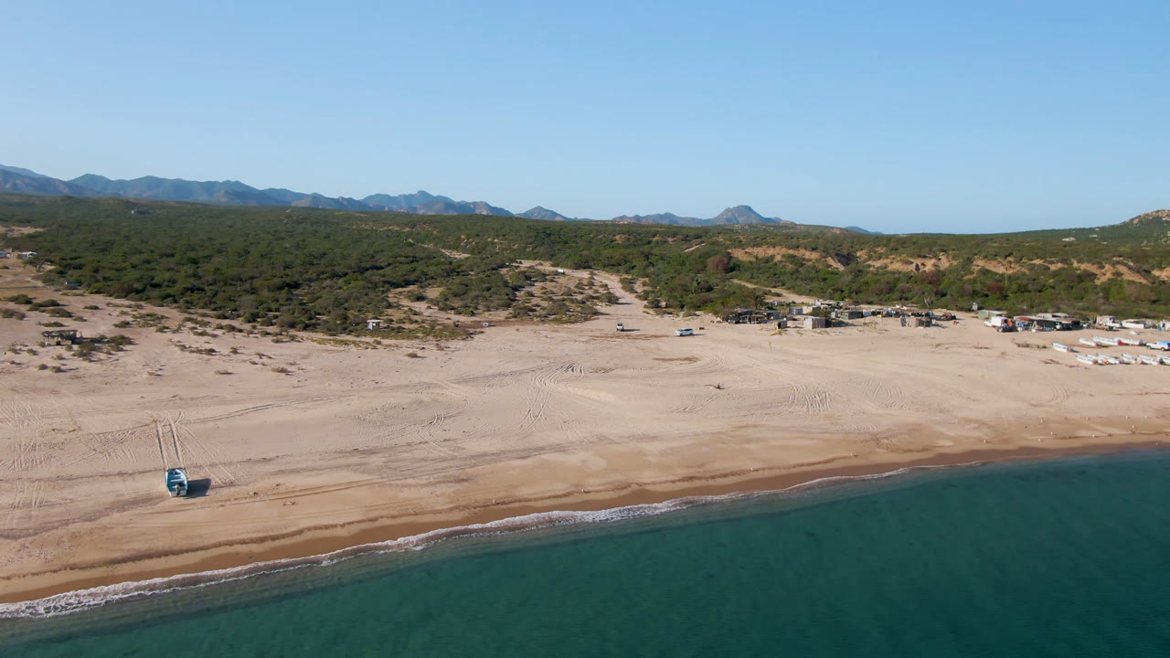 Aerial view in Los Frailes showing the shoreline across the lower frame, tire-marked sand, low green vegetation, distant hills, and no visible people.