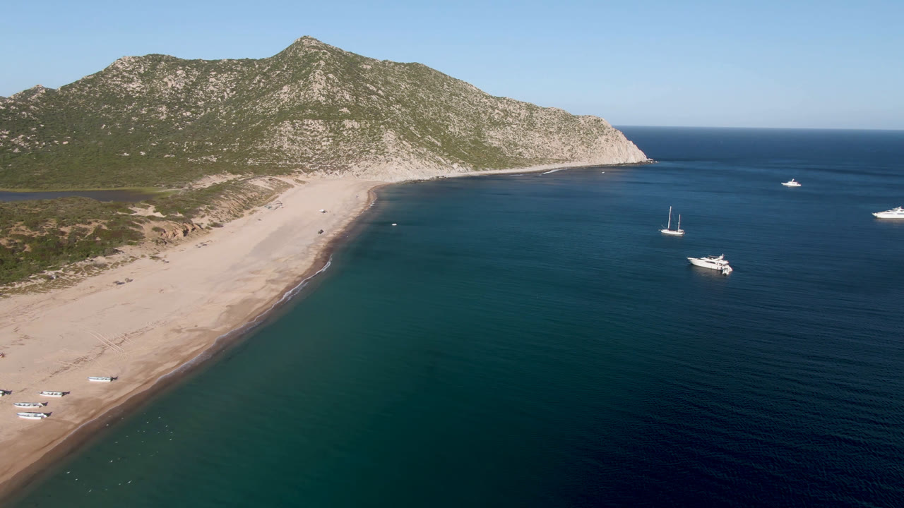 High aerial view over Los Frailes, Baja California Sur, with a long sandy beach, rocky green headland, several boats offshore, and no visible people.