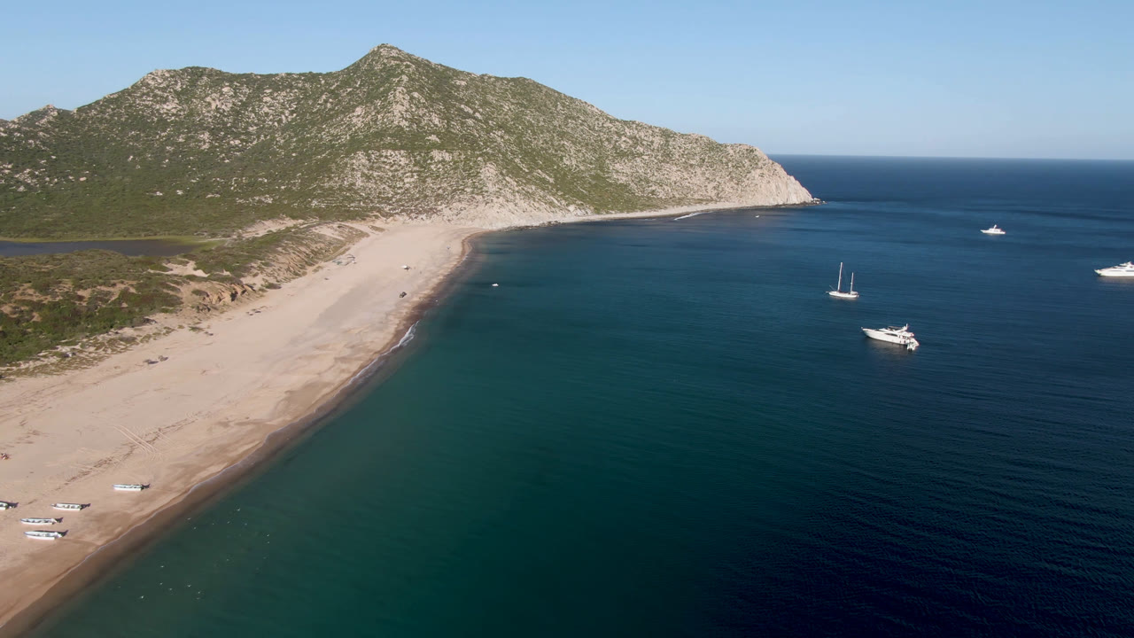 Aerial frame at Los Frailes showing the curved sandbar edge, teal shallows, sailboats and a motorboat offshore, with no visible people on the beach or boats.