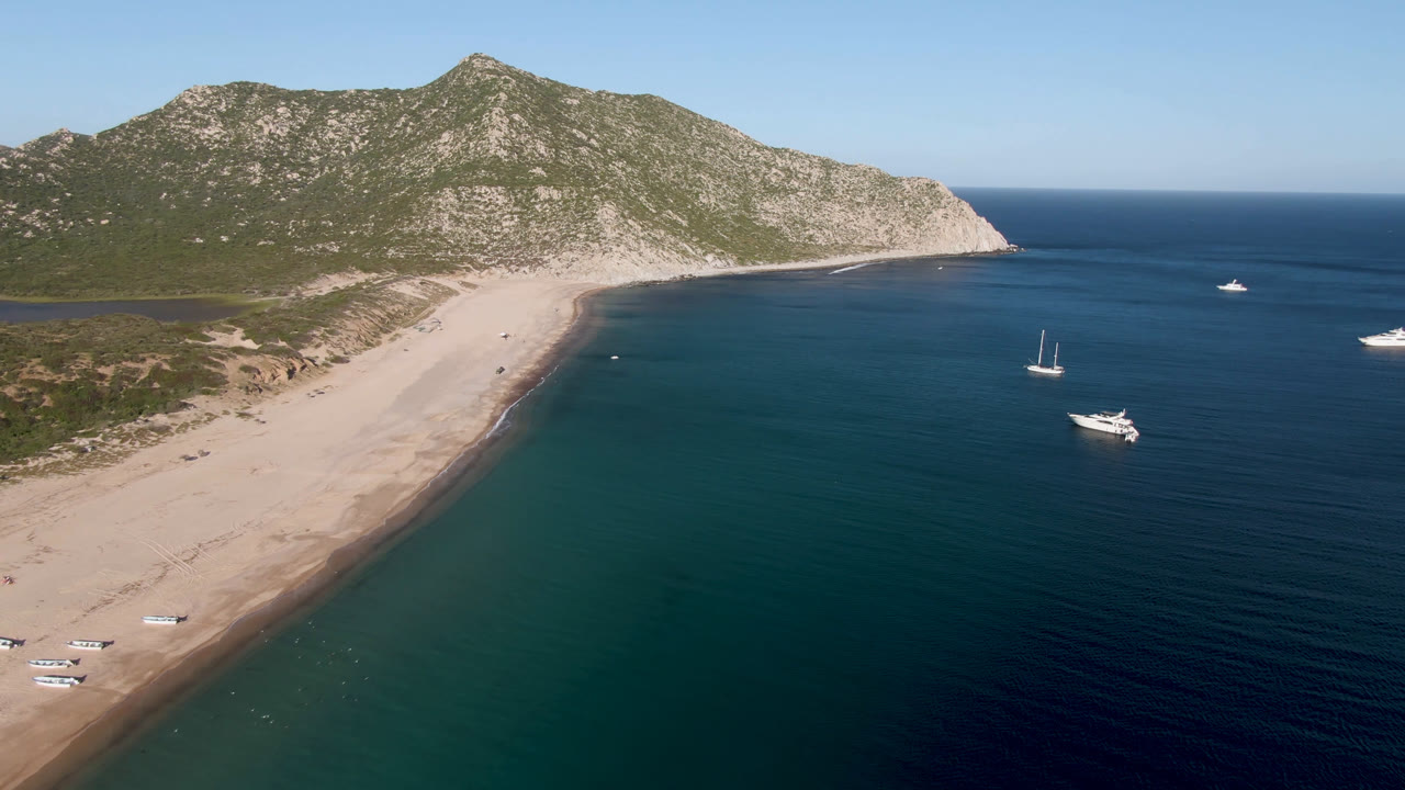 Wide coastal aerial with pale beach, rocky point, calm ocean, and boats spread offshore.