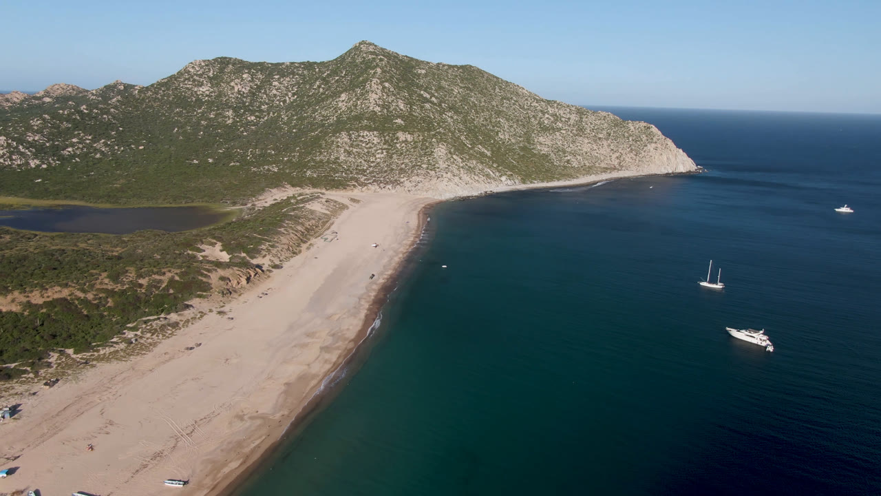 Aerial frame over Los Frailes, Baja California Sur, with a curved sandy beach at left, several boats offshore at right, rocky green hills beyond, and no visible people.