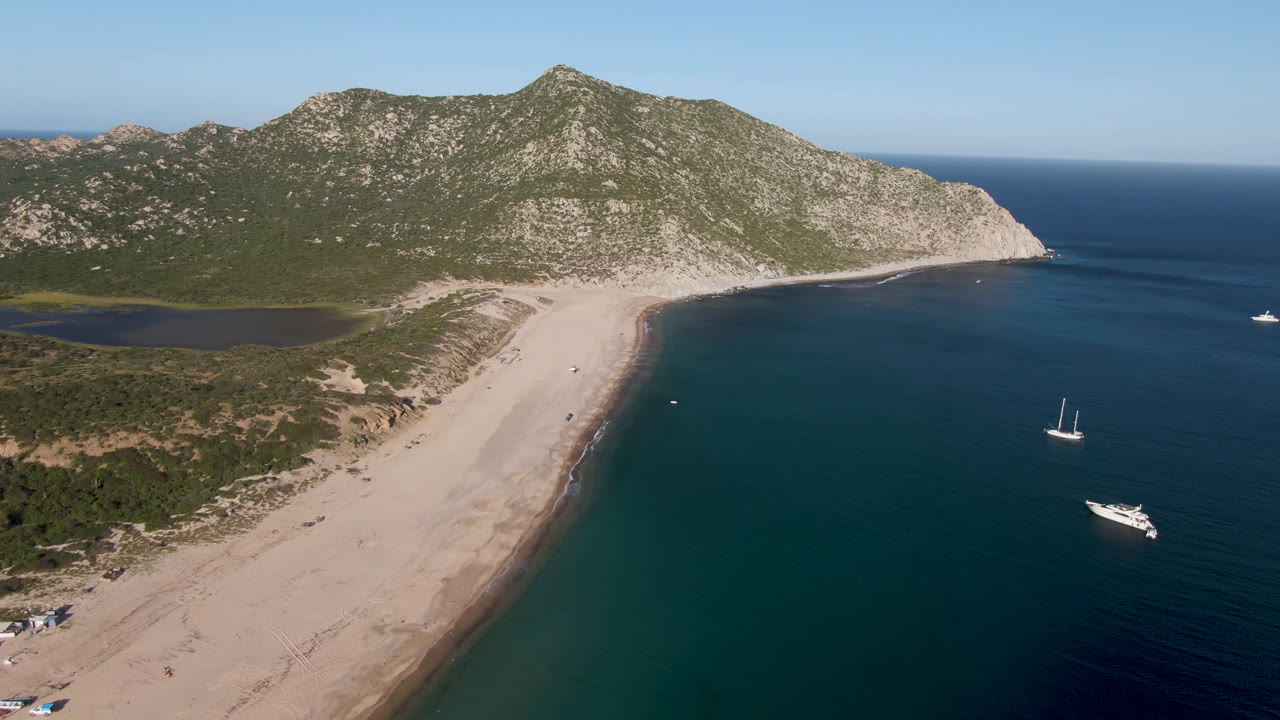 Wide aerial coastline with empty sand, offshore boats, and a mountain ridge.