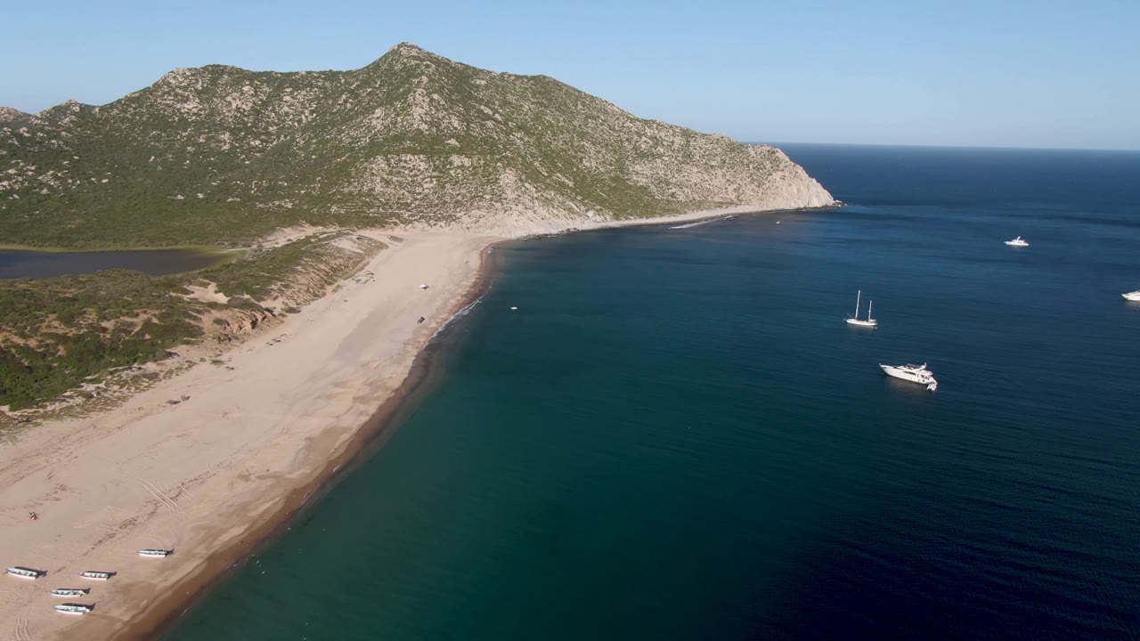 Opening aerial frame over Los Frailes, Baja California Sur, Mexico, with a long sandy beach, several small boats offshore, rocky green hills behind, and no visible people.