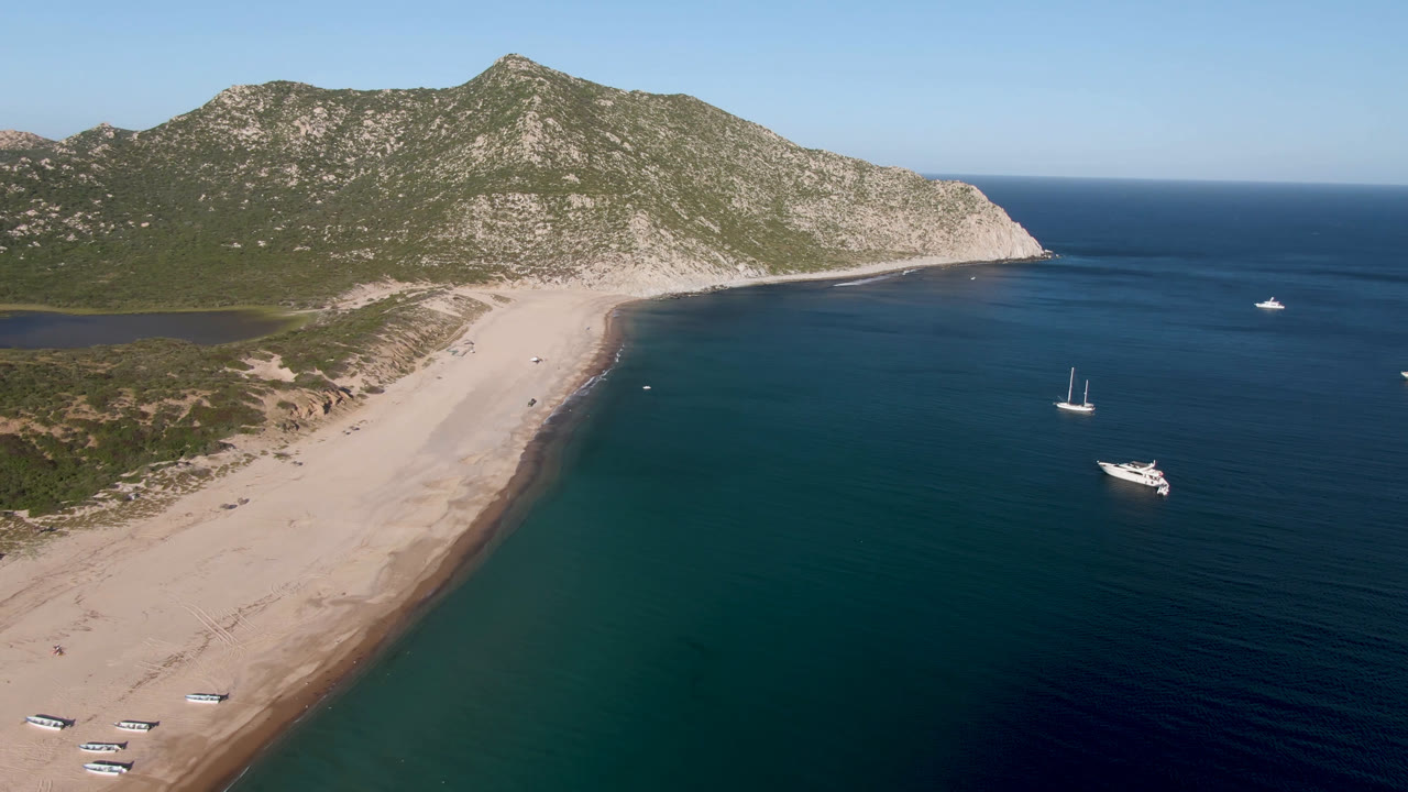 Second frame over Los Frailes shows the sandy beach angled along the left, anchored sailboats and a larger motorboat in blue water, hillside terrain, and no visible people.