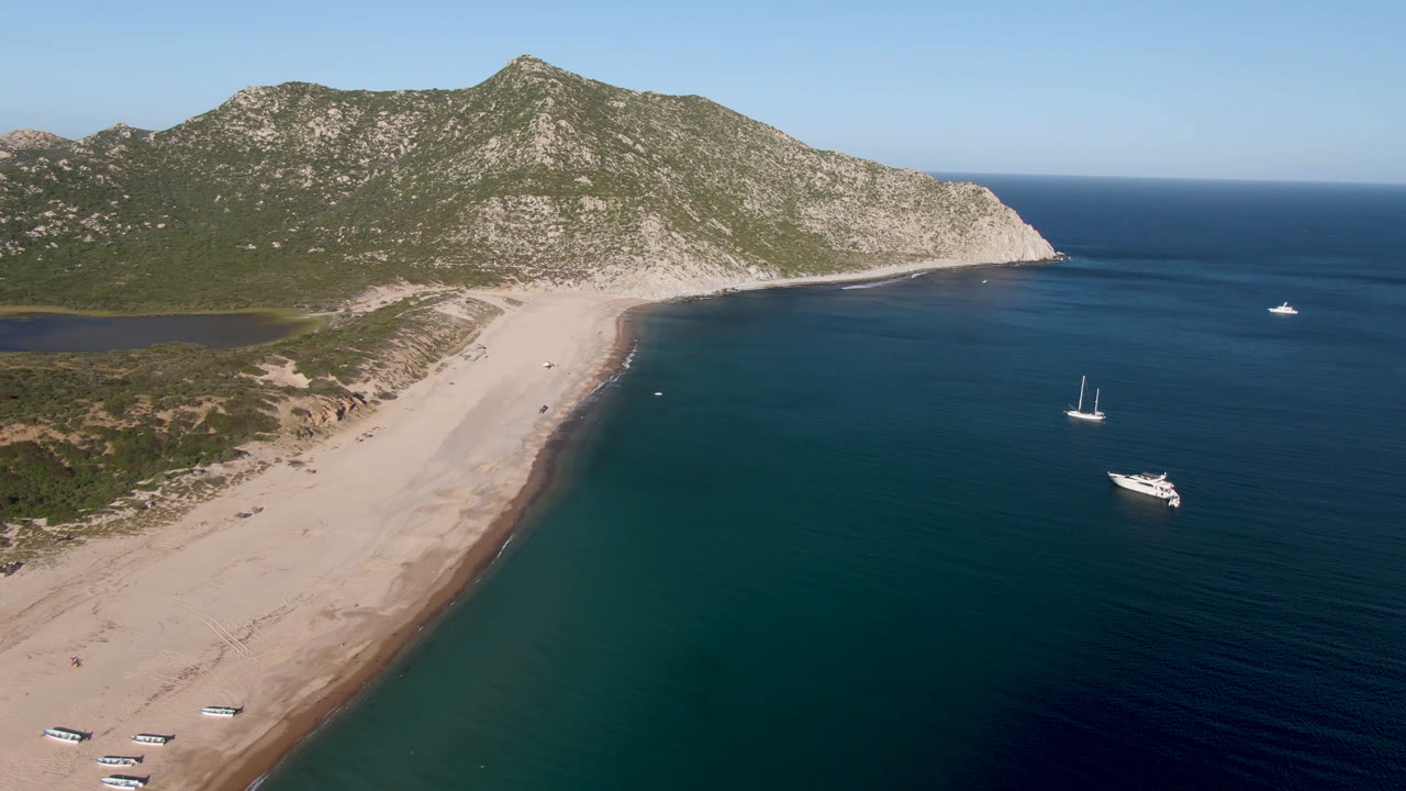 Drone view of a beach bay with sailboats, a motorboat, and rocky hills under clear sky.