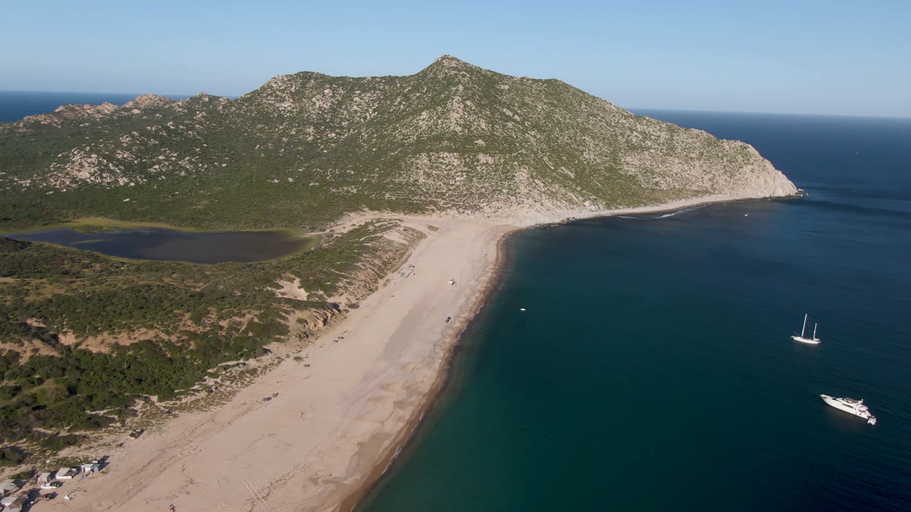 A high aerial view over Los Frailes shows a pale sandy beach curving between green rocky hills and blue ocean, with a sailboat and motor yacht offshore and no visible people.