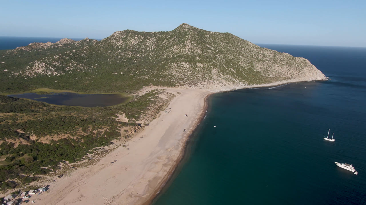 At 0:01, the Los Frailes aerial keeps the beach centered between the scrub-covered hills and open water, with the sailboat and motor yacht still visible offshore and no visible people.