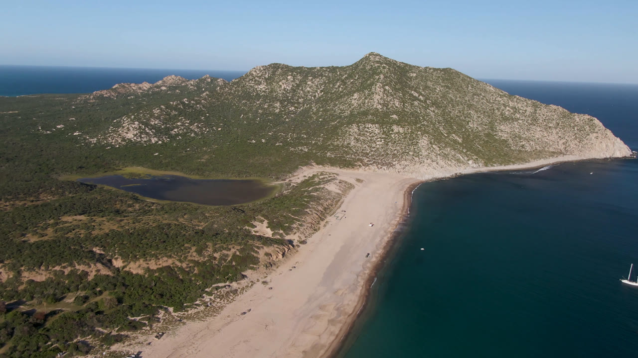 Aerial view at Los Frailes with the beach running diagonally beside the ocean, a dark inland water area behind the dunes, rocky green-brown hills, and no visible people.