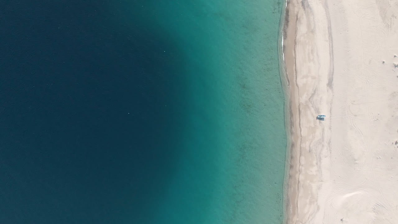 Aerial frame over Los Frailes showing a vertical strip of pale sand, blue-green water on the left, a small blue boat set back from the surf, and no visible people.