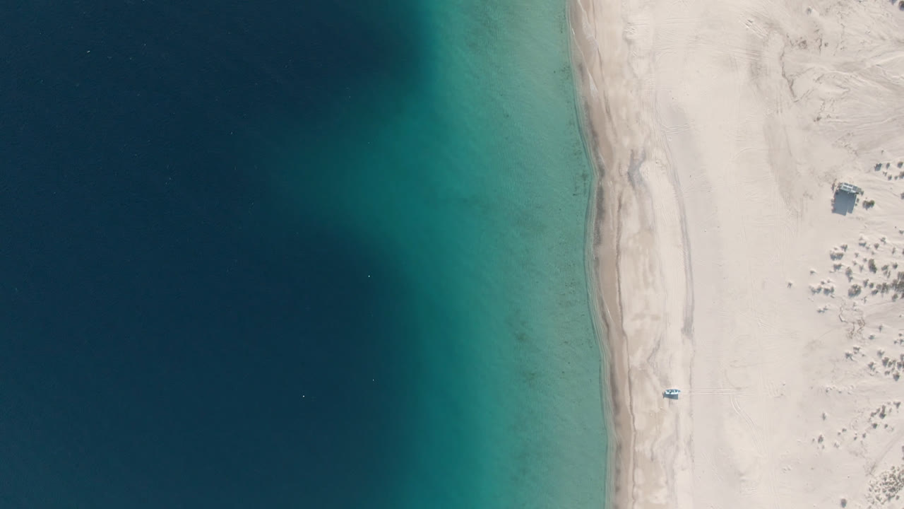 Top-down aerial frame of Los Frailes with deep blue water at left, turquoise shallows along the shoreline, pale sand at right, a small boat on the beach, and no visible people.