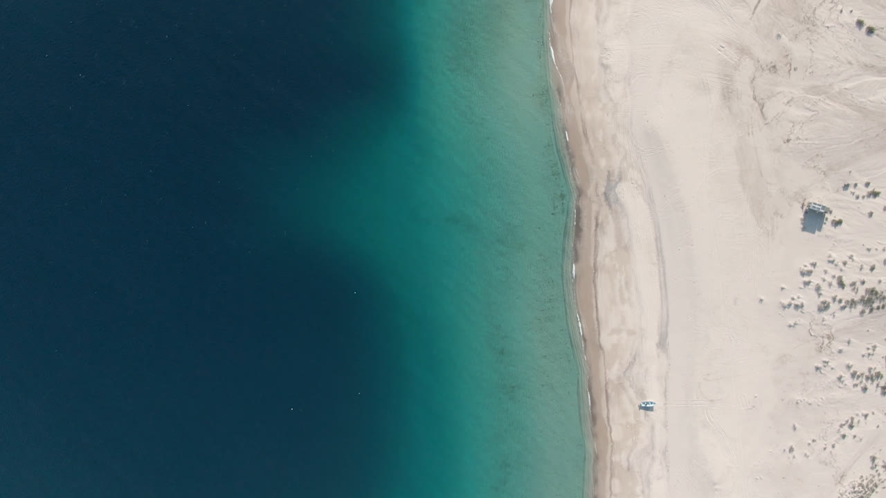 Aerial view over Los Frailes showing a vertical strip of beach between dark water and light desert sand, with the small beach boat slightly lower in frame and no visible people.